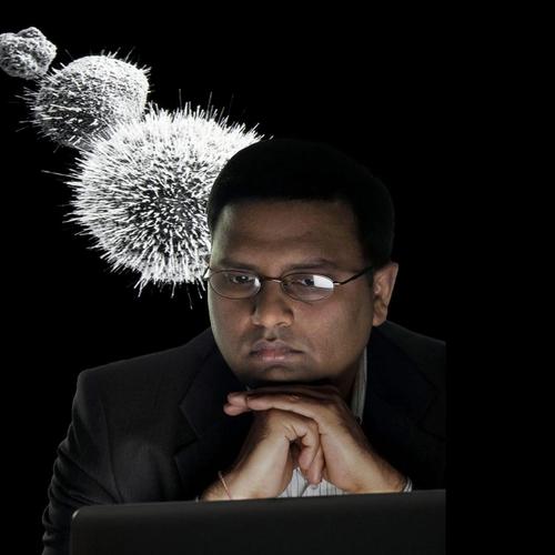 A serious-looking man with glasses and a suit sits in front of a laptop, with his chin resting on his clasped hands, while three spiky microscopic objects float above his head against a black background.